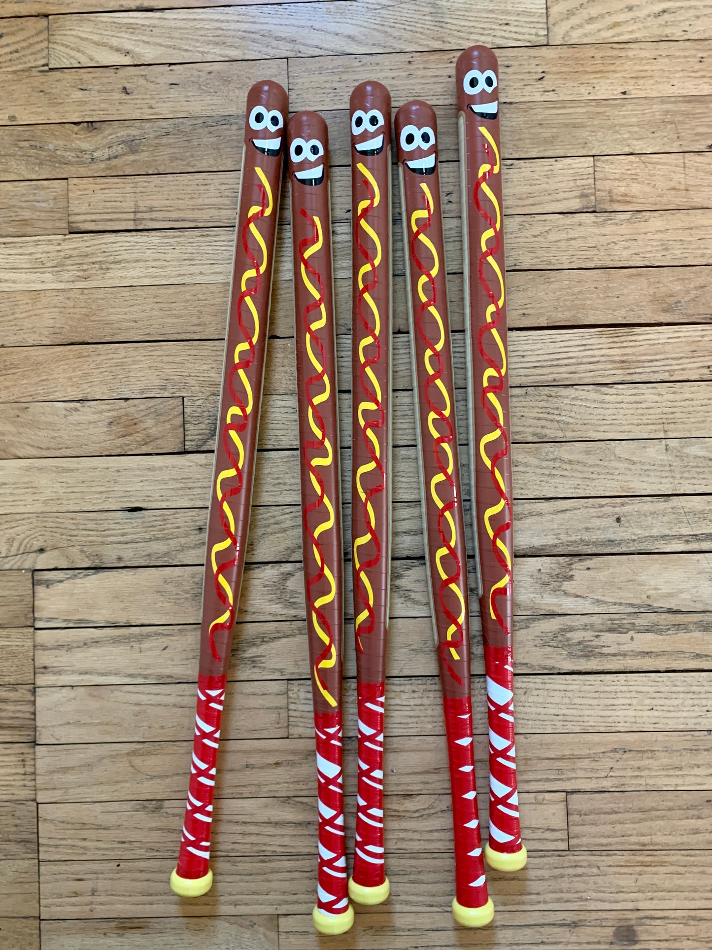Set of five custom hot dog design WIFFLE bats with smiling faces and mustard graphics on wood floor