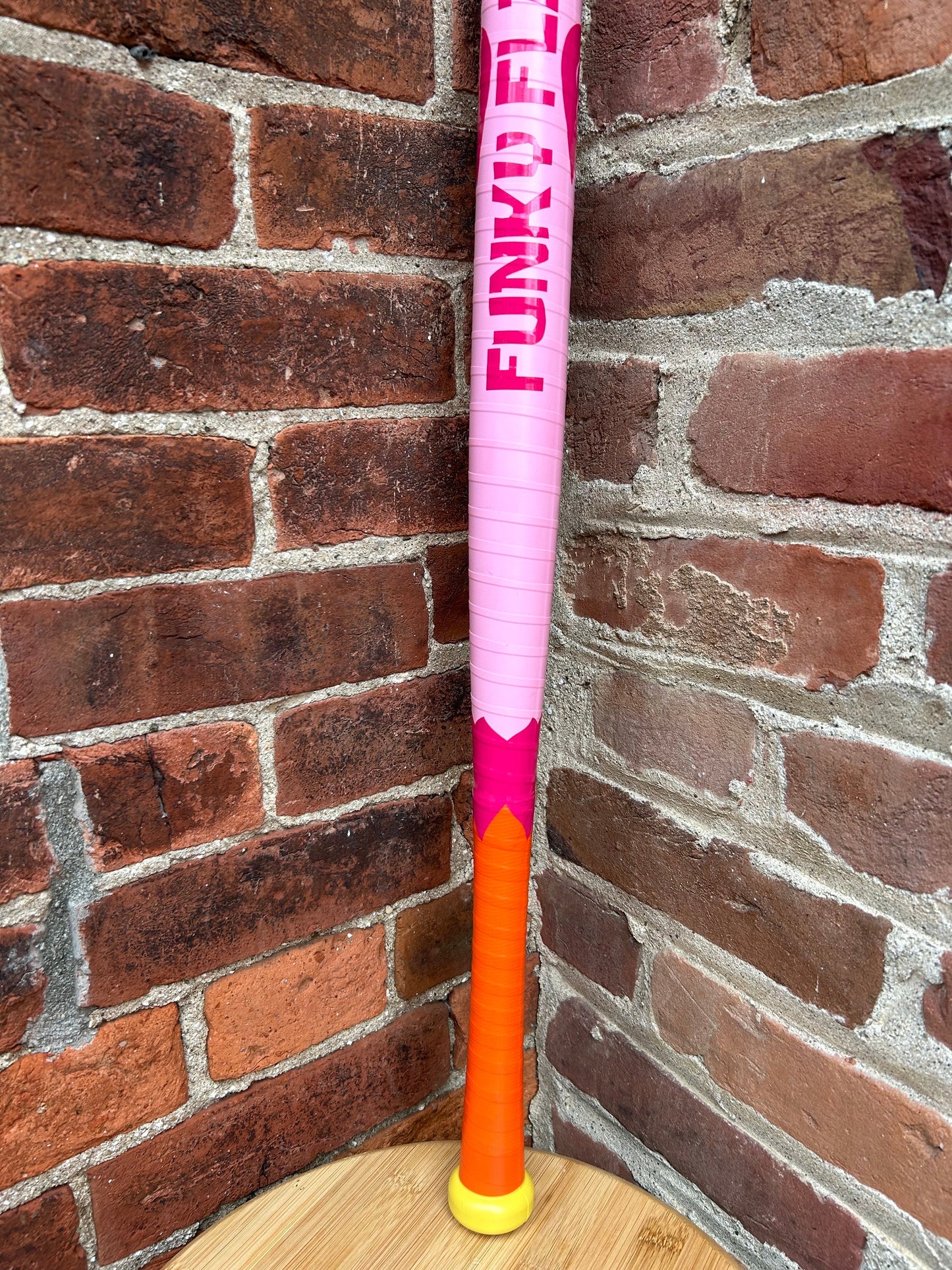 Custom pink and orange Wiffle bat with FUNKY FELT text, leaning against a brick wall.